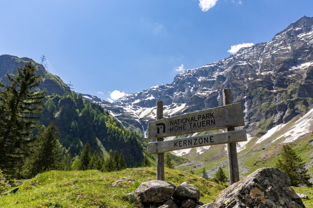 Image of Hohe Tauern National Park in Austria showing mountains, clear blue sky and a sign welcoming people to the park.