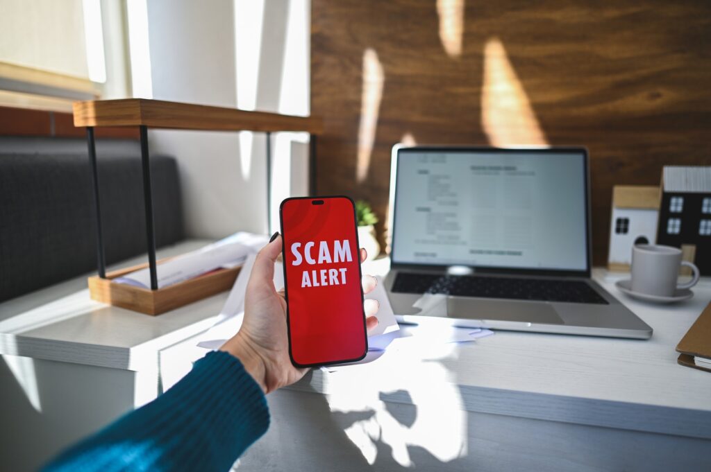 A home office pictured with a working laptop open and red scam alert visible on a nearby mobile phone.