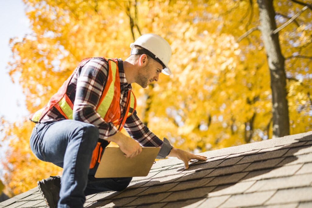 A man in a hard hat and high vis vest inspecting a roof of a home for damage. Bright orange autumn trees in the background.