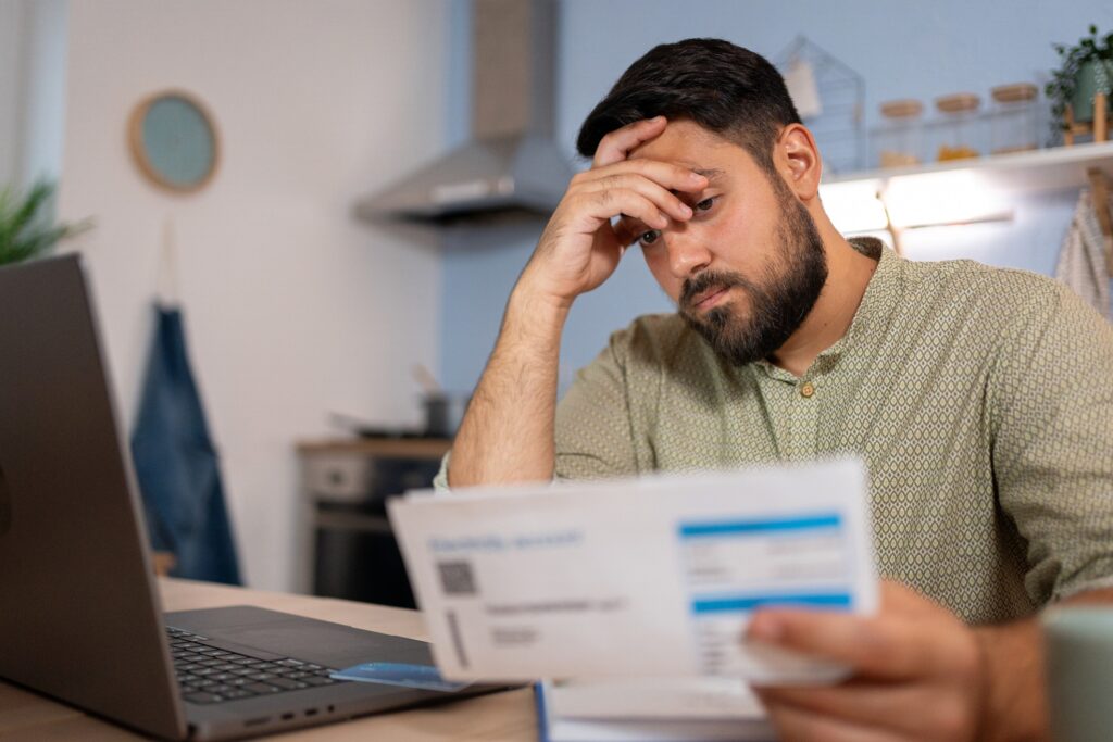 A man in his modern kitchen in front of a laptop looking confusion about a bill.