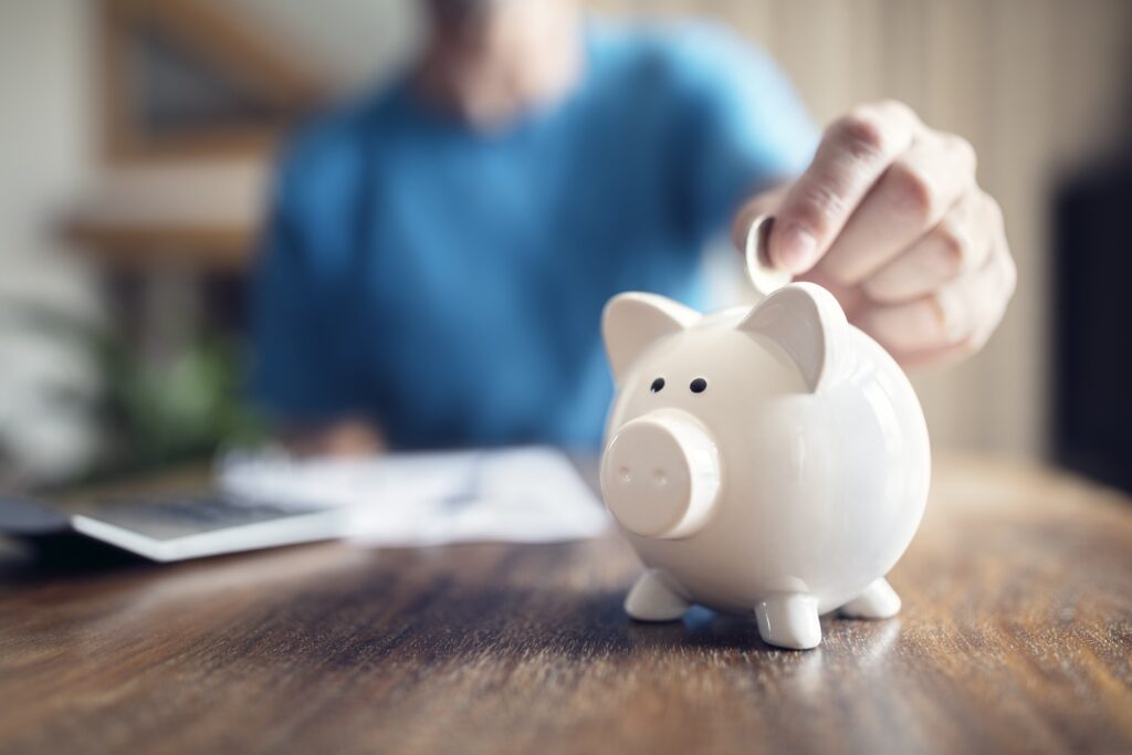 A person in a blue jumper is reviewing their finances and putting some savings into a white piggybank - receipts and a calculator are also visible.