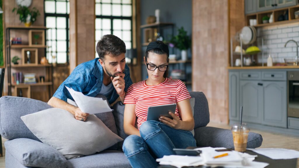 A couple sit reviewing bills in their modern apartment, with paperwork, a tablet and a calculator visible.