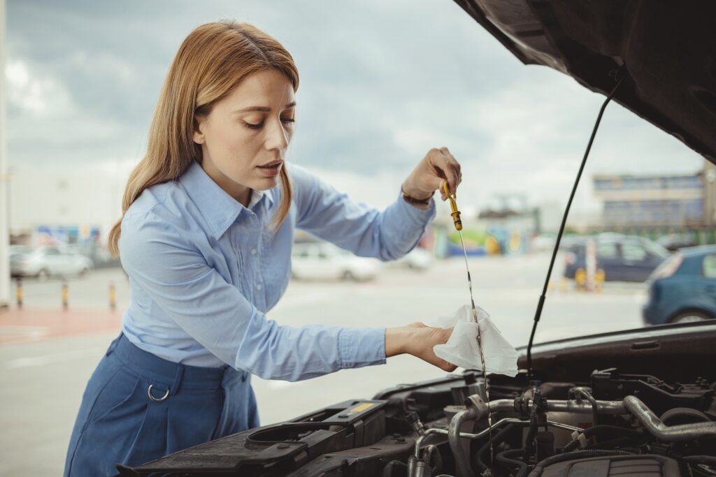 A woman dressed in a blue shirt and trousers checking the oil on her car with the engine visible and the bonnet open.