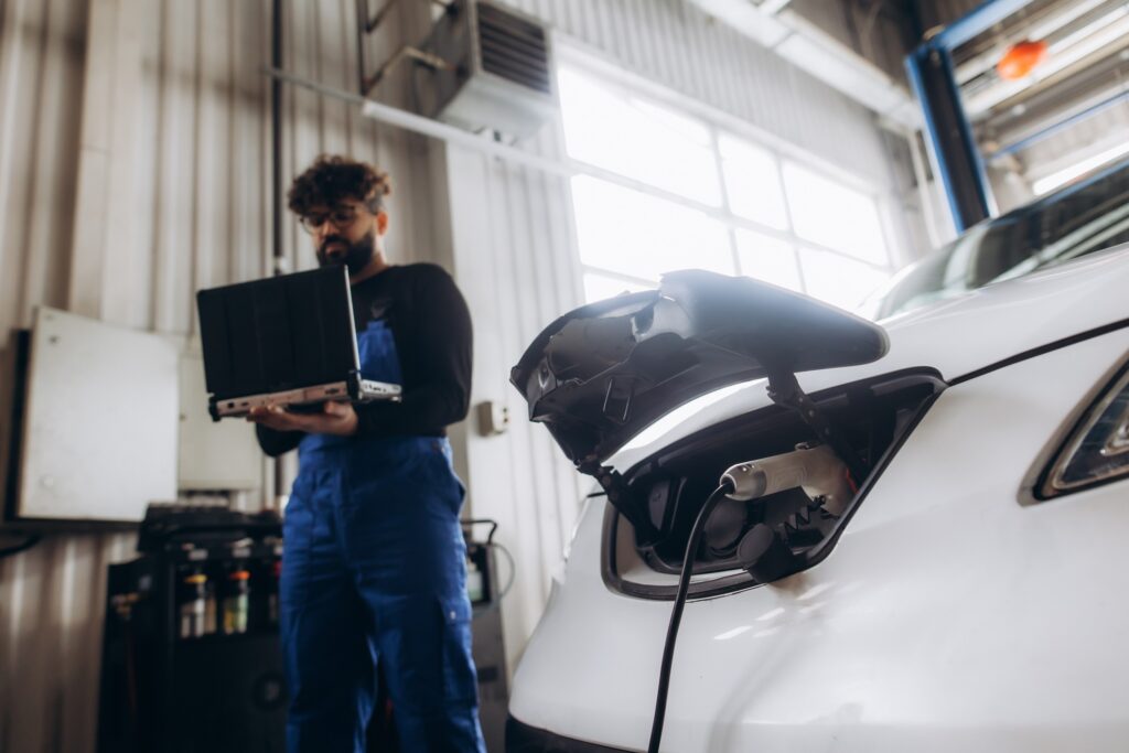 A mechanic fixing a white electric vehicle in his garage, charging cable visible.